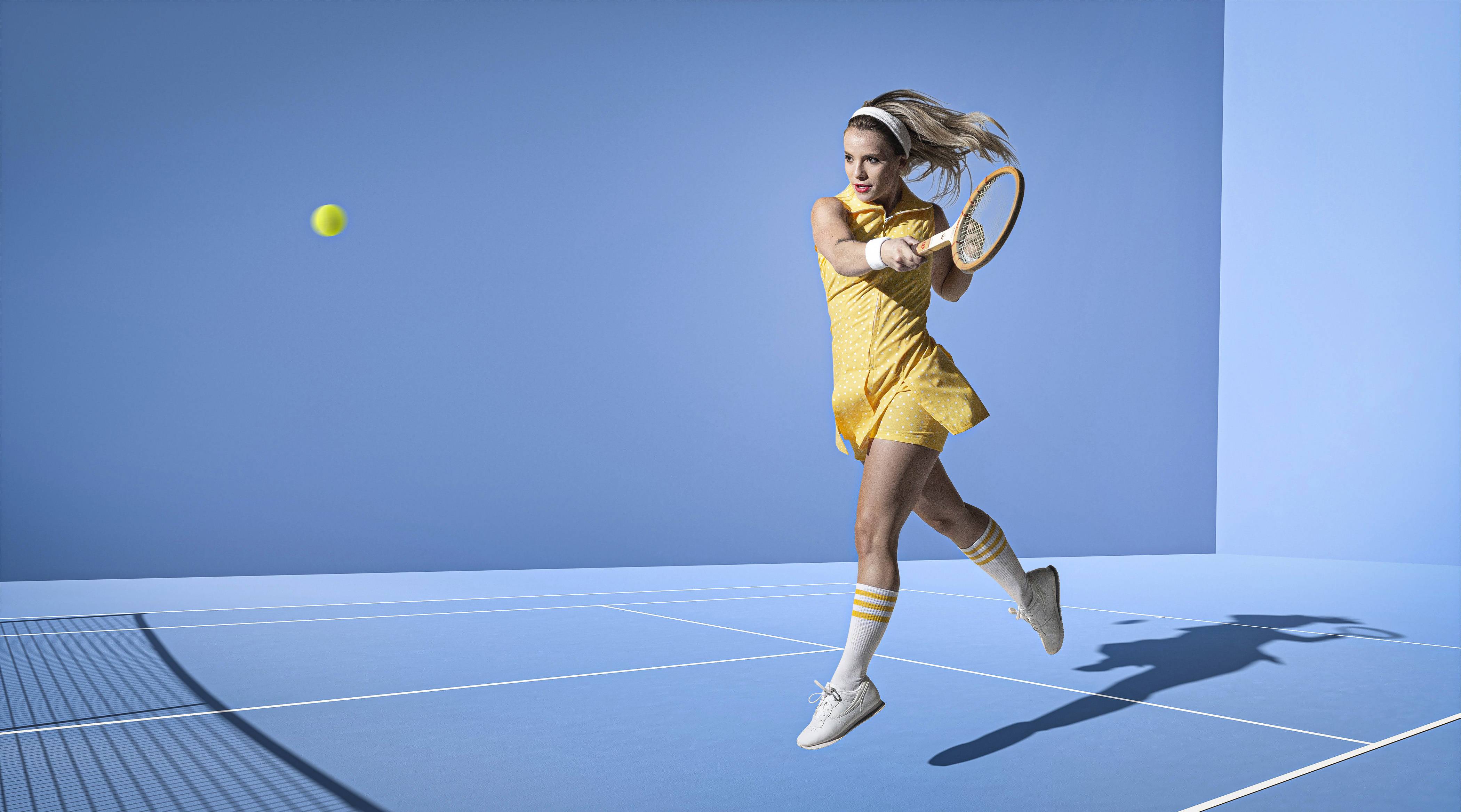 Female model hitting a tennis ball in peak action on a blue studio court lit by a single light mimicking sunlight