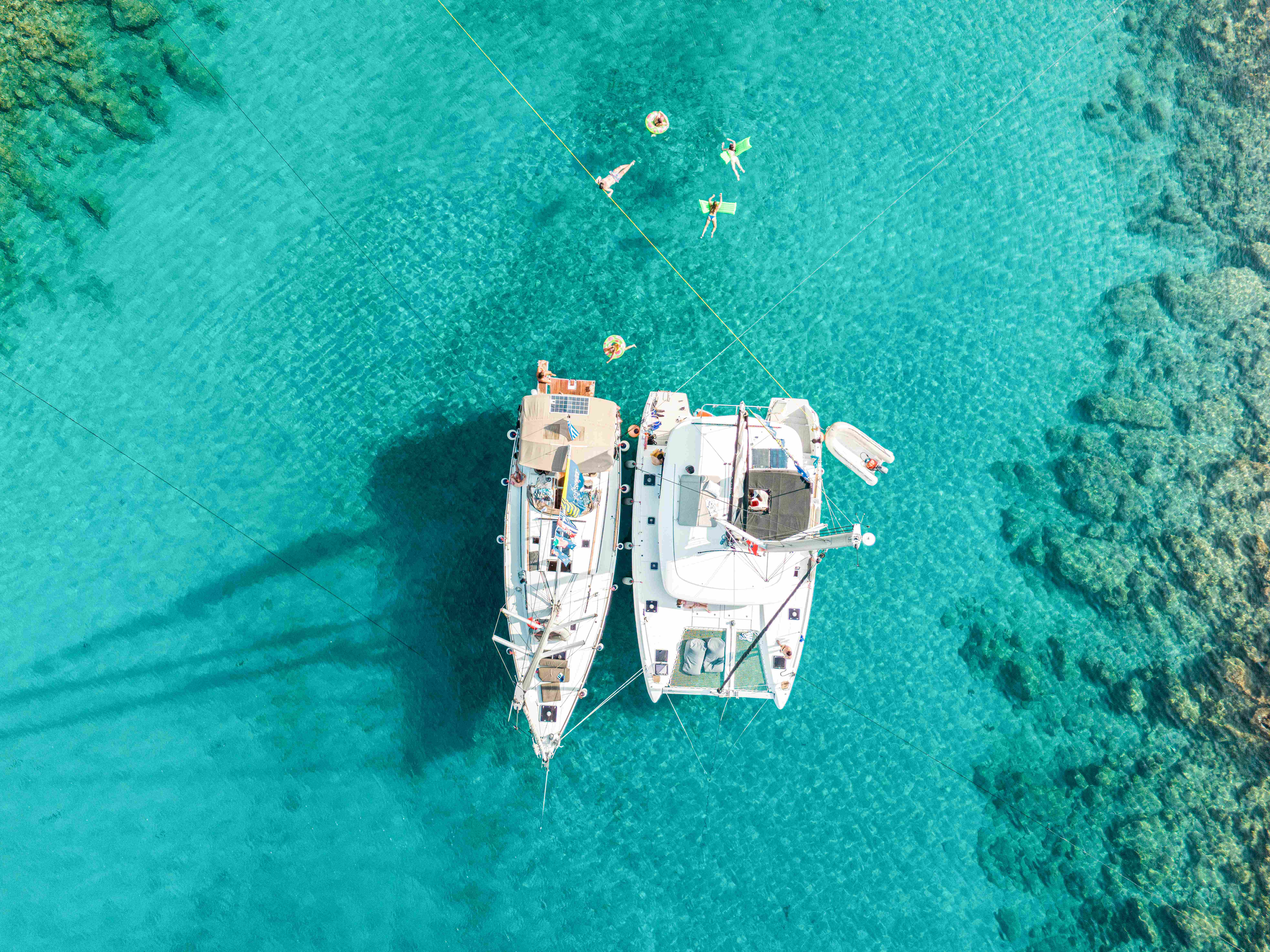 Aerial view of a catamaran and sailboat with people relaxing in turquoise waters in Greece